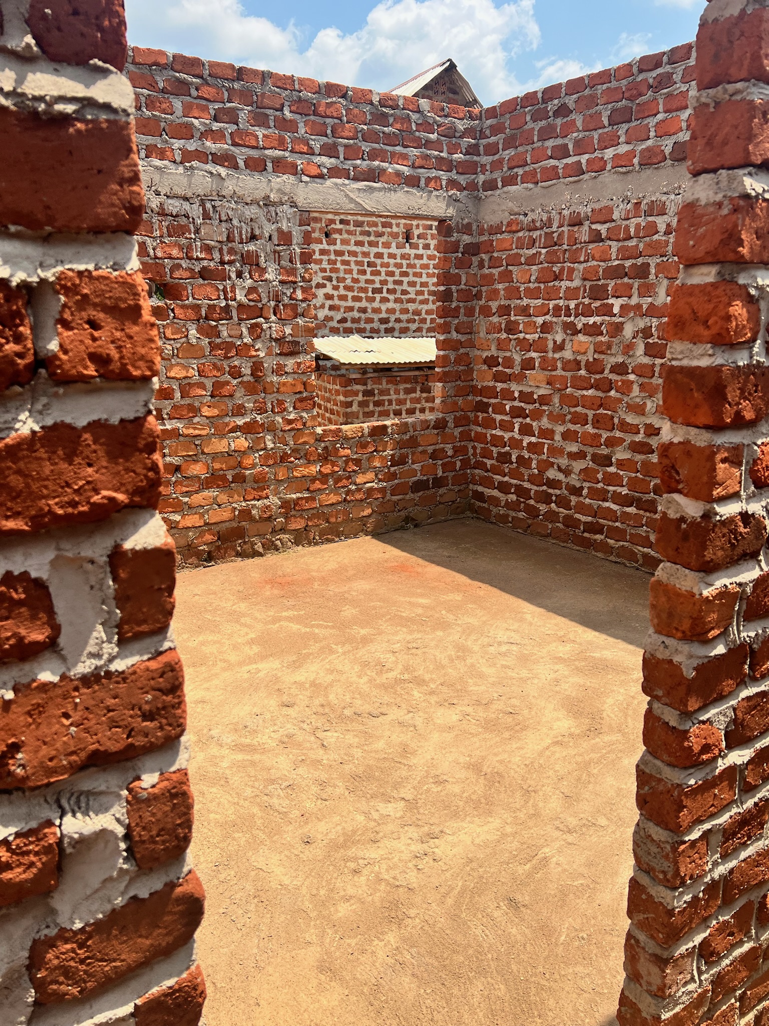 Unroofed dormitory room with raw brick walls and a dirt floor.