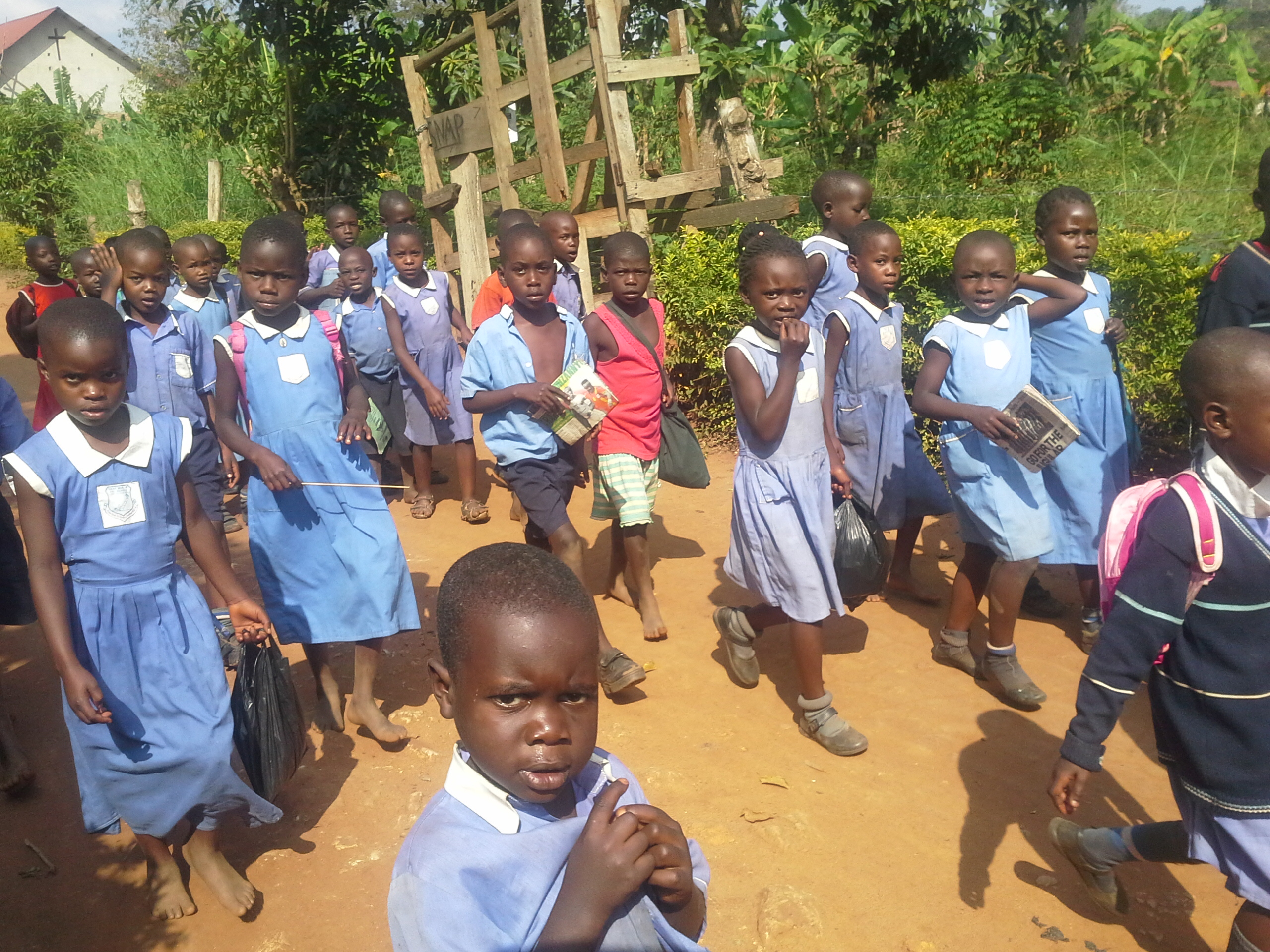 Children in blue SNAP school uniforms walking a dirt path past a wooden SNAP sign, July 2017.